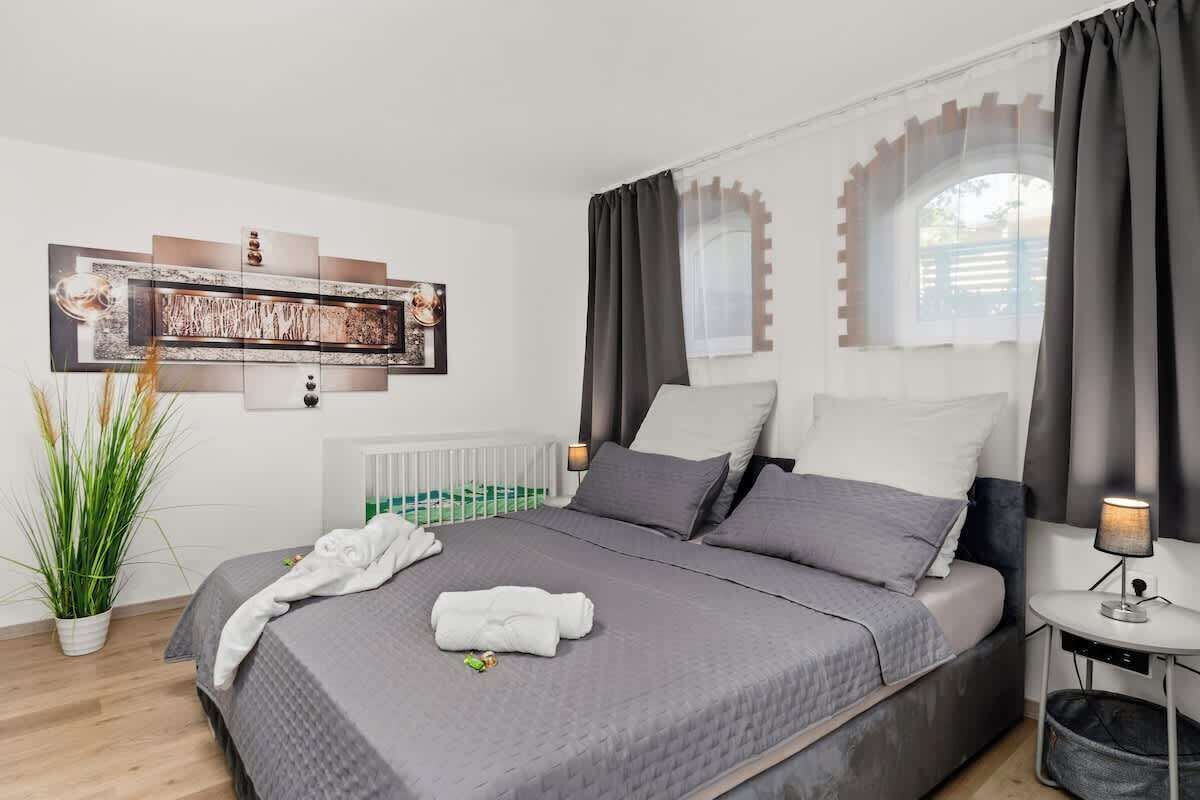 A modern hotel room with a large bed, grey bedding, a small bedside table with a lamp, a potted plant, and a wall-mounted artwork.