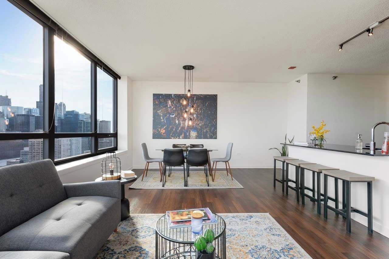 A modern living room with a grey sofa, a dining area with a table and chairs, a kitchen with bar stools, and a large window with a city view.