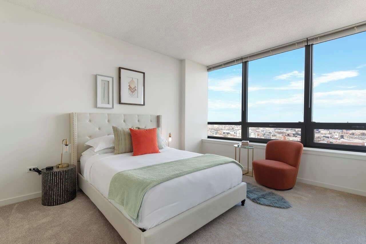 Une chambre moderne avec un grand lit, un fauteuil rouge et une vue sur la ville à travers de grandes fenêtres.