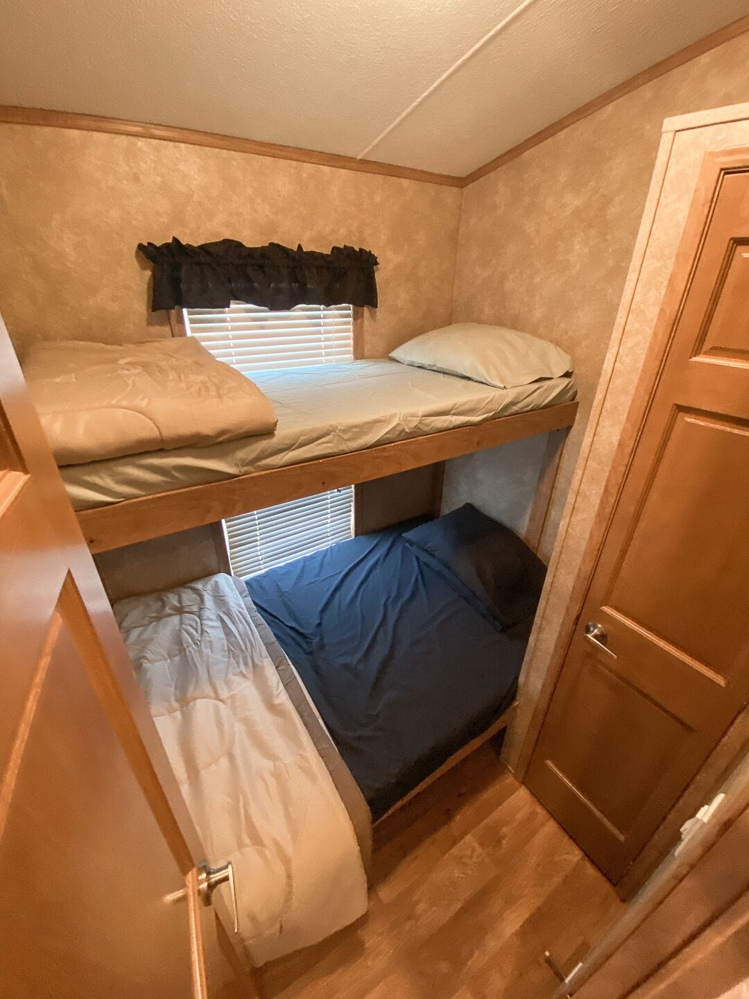A compact RV interior with a bunk bed, a small window with blinds, and a wooden door.