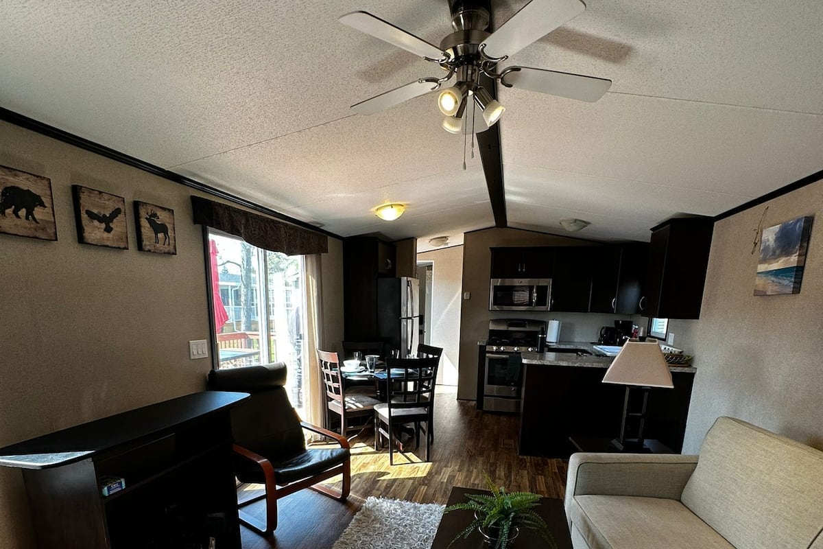 A modern living room with a sofa, coffee table, and a ceiling fan. There is a kitchen area with a microwave and stove in the background.