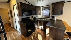 A compact kitchen with dark wood cabinets, a black stove, and a window with blinds. - Falcon Cottages (Niagara-on-the-Lake)