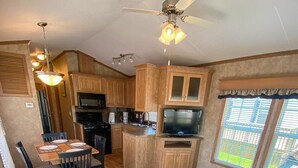 A compact RV kitchen with wooden cabinets, a dining area with chairs, a gray sofa, and a ceiling fan. - Falcon Cottages (Niagara-on-the-Lake)