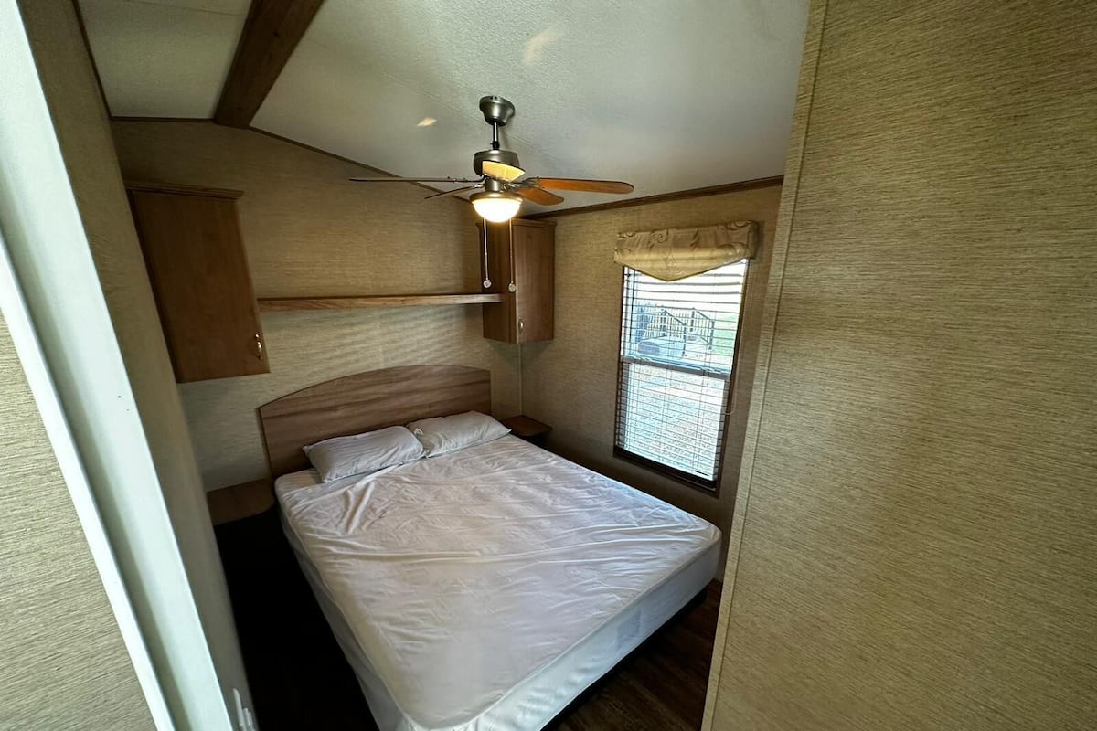 A bedroom with a bed, a ceiling fan, a window with blinds, and wooden walls.