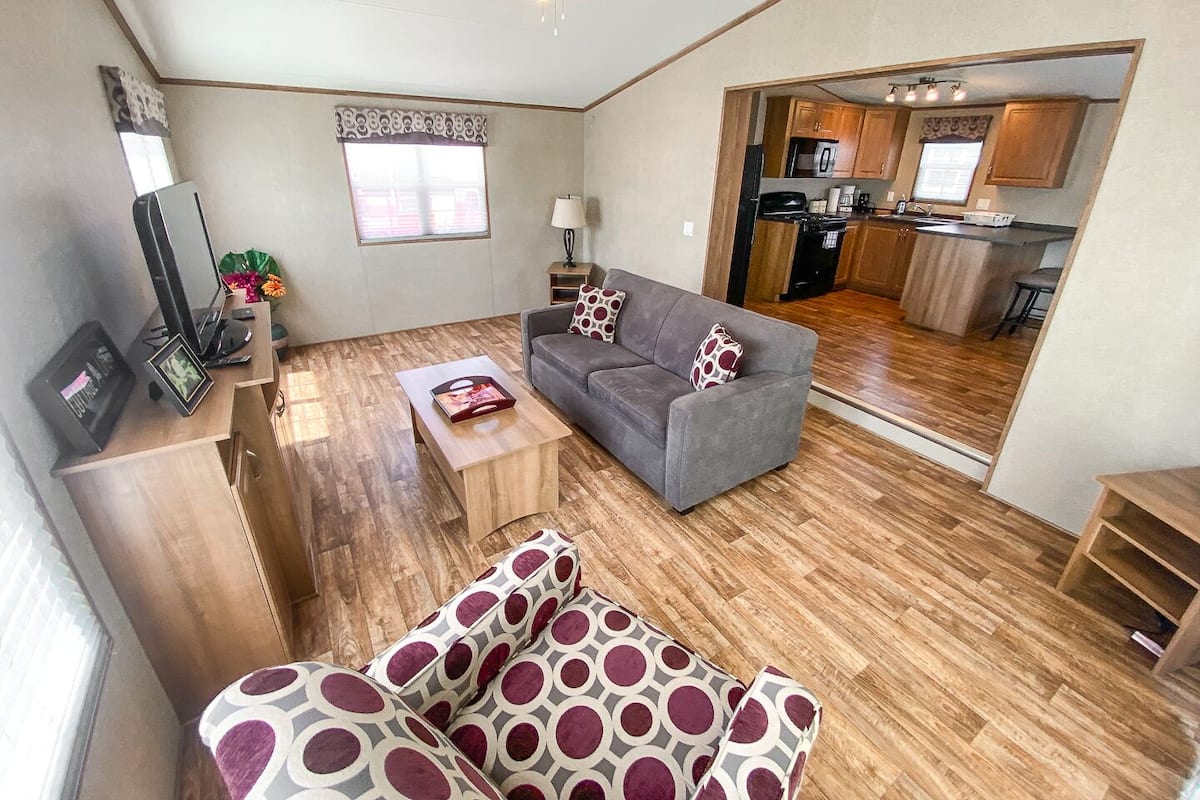 A compact living space with a grey sofa, patterned cushions, a wooden coffee table, and a TV stand. A ceiling fan is visible, and the room features wooden flooring.