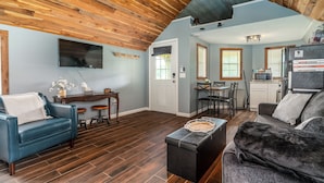 Una sala de estar con un sofá azul, un sofá gris, techo de madera, televisor de pantalla plana y una zona de cocina con refrigerador y microondas.