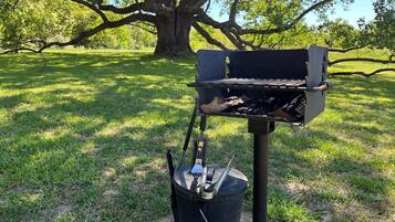 Una parrilla portátil con una olla y utensilios, sobre un área cubierta de césped con un gran árbol al fondo.