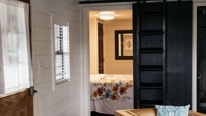 Ein Zimmer mit einem Bett, einer Holztür, einem Fenster mit Jalousien und einem dunklen Holzboden.