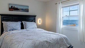 A bedroom with a bed, a nightstand, a wall-mounted lamp, a window with curtains, and a painting on the wall.
