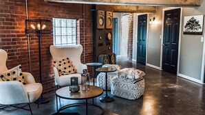 Un salon moderne avec un mur en briques, une table basse ronde, deux fauteuils blancs à pois noirs, une petite table ronde avec une lampe et un pouf en osier.