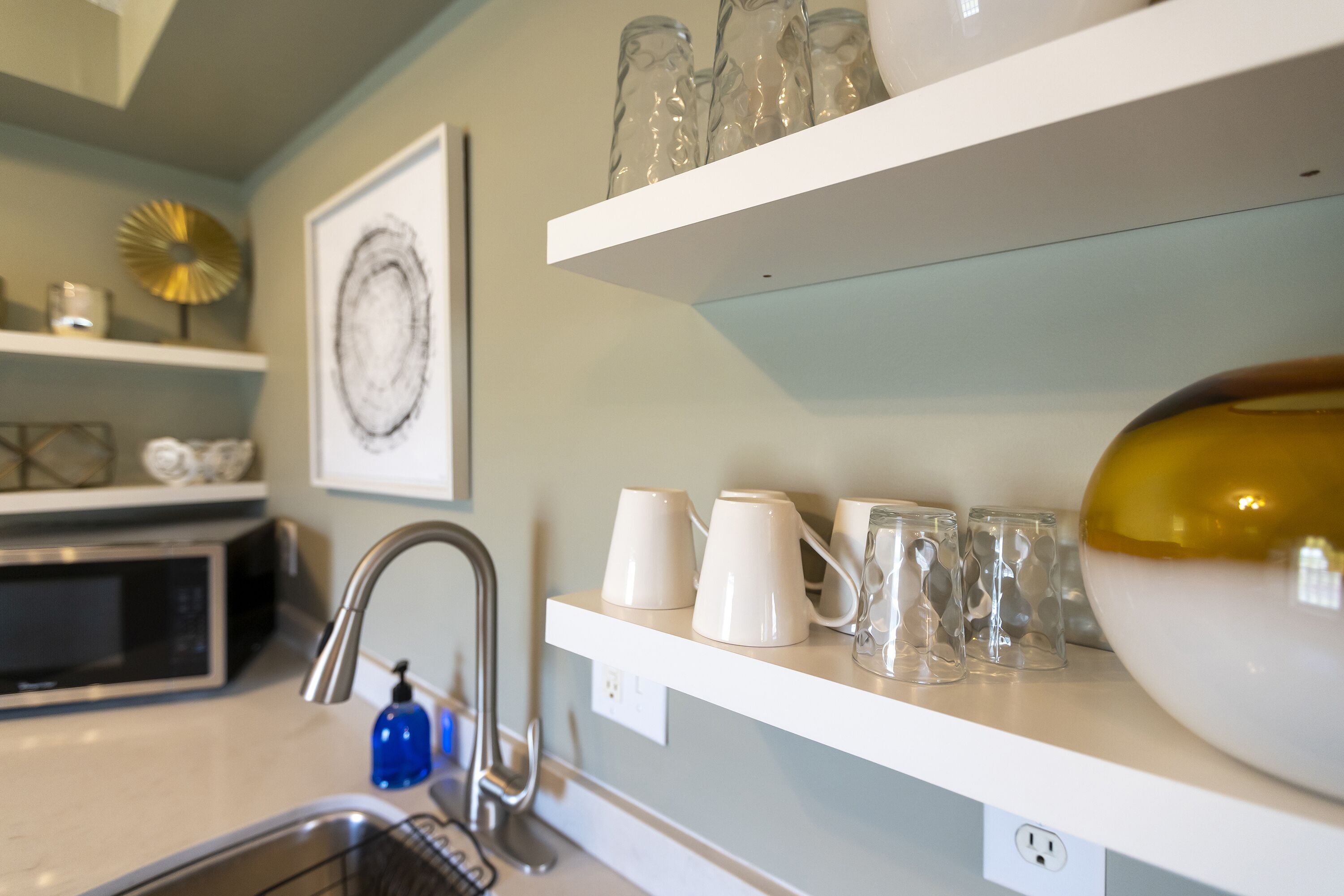 A kitchen with white shelves, a microwave, a sink, and a blue soap dispenser.