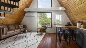 A loft-style living room with a wooden ceiling, a large window, a sofa, a chair, and a kitchen area with bar stools. - 2BR A-Frame Cabin w/ Hot Tub Near Wine Trail (Makanda)