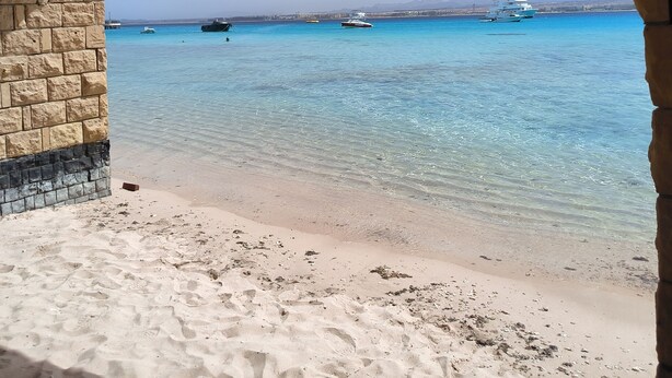 A beach view through a stone archway with clear turquoise water, boats, and distant mountains.