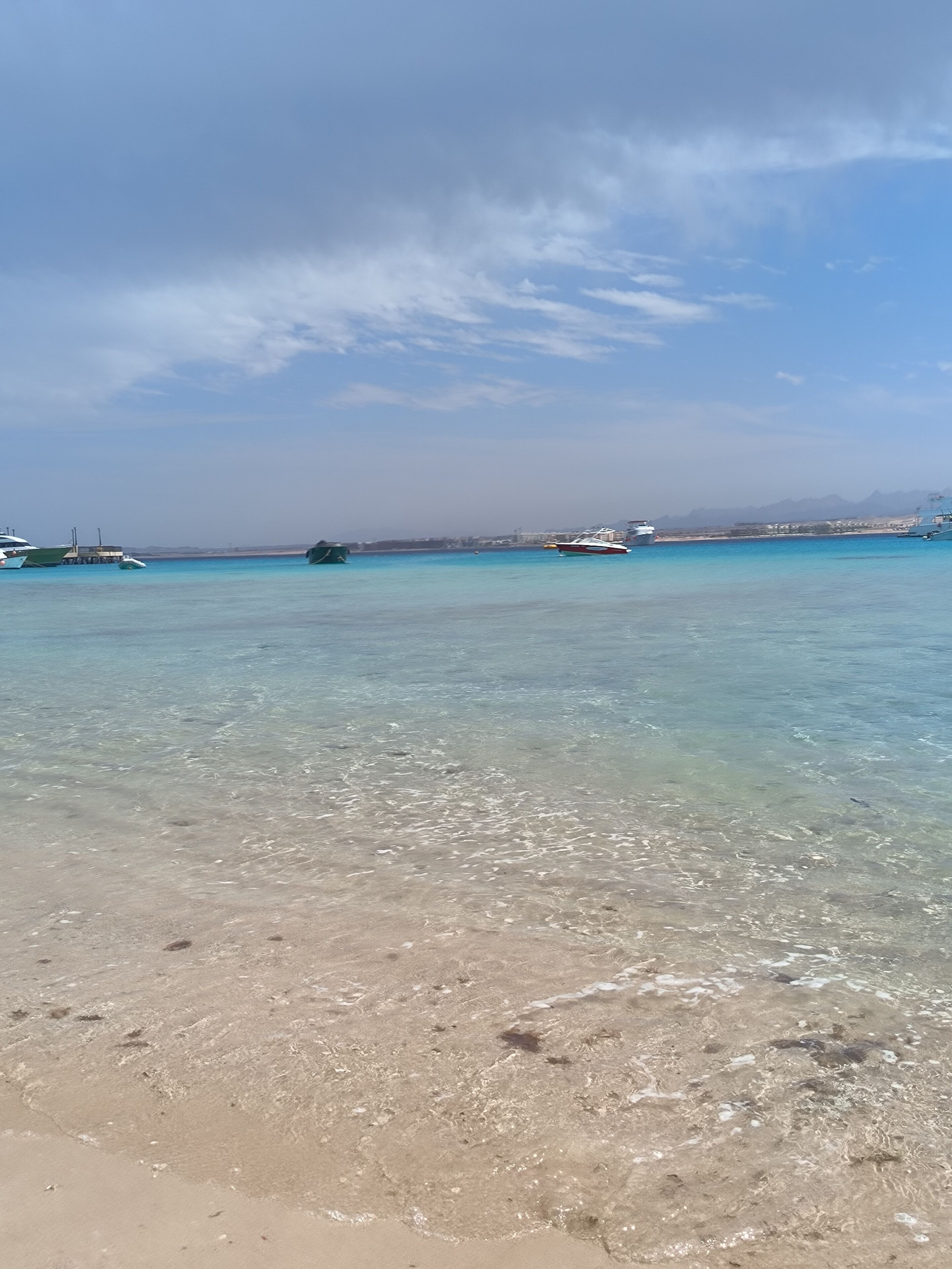 Una playa con aguas turquesas cristalinas, barcos y montañas a lo lejos.