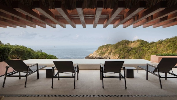 A terrace with four chairs and a table, overlooking a coastal landscape with a view of the sea and a lighthouse.