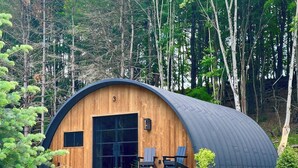 Une cabane en bois dotée d’un toit incurvé, de grandes fenêtres et d’un petit espace patio aménagé avec des sièges.