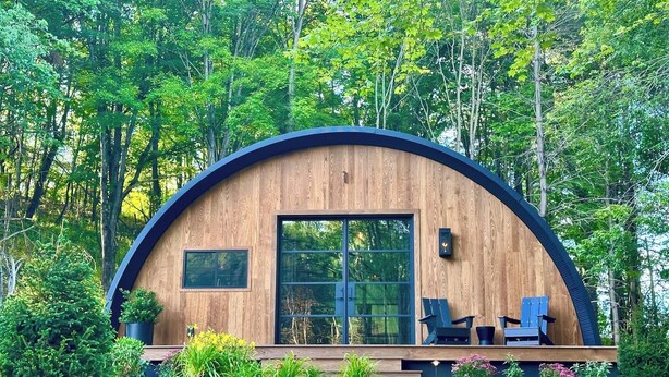Une cabane moderne en bois, de forme semi-circulaire, dotée de grandes fenêtres et entourée d’une végétation luxuriante et d’arbres.