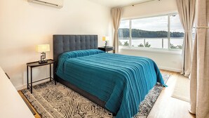 A bedroom with a large bed, a nightstand, a lamp, a window with a view of a lake, and an air conditioning unit. - The Cliffside Poustinia (Kodiak)