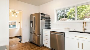 A modern kitchen with stainless steel appliances, white cabinetry, and a window providing a view of greenery outside. - The Cliffside Poustinia (Kodiak)