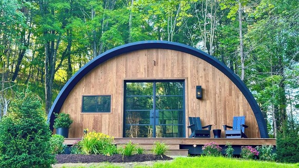 A modern, semi-circular wooden cabin with large windows, surrounded by lush greenery. - Treat yourself at the stunning June Farms Wellness Huts! (West Sand Lake)