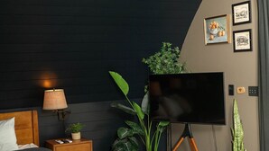 A modern bedroom with a large bed, a flat-screen TV on a tripod stand, a bedside table with a lamp, and several potted plants. - Treat yourself at the stunning June Farms Wellness Huts! (West Sand Lake)