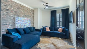 A living room with a sectional sofa, a ceiling fan, a brick wall, and a large window with curtains.