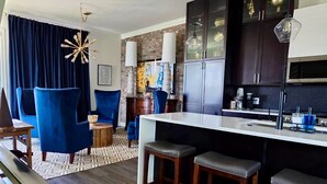 A modern kitchen with dark cabinets, a white countertop, and bar stools. There is a dining area with blue chairs and a patterned rug.