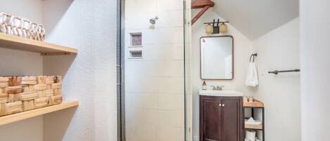 A narrow bathroom with a shower, a wooden shelf with baskets, a mirror, and a small cabinet.