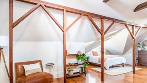 A bedroom with a sloped ceiling, wooden beams, a bed, a chair, a small table, and a potted plant. - Sac City Loft (Sacramento)