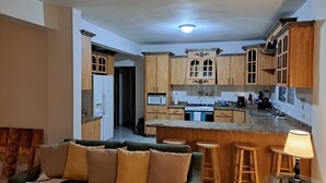 Una cocina moderna con armarios de madera, microondas empotrado y cocina a gas. Cuenta con una zona de bar con taburetes y una pequeña mesa con lámpara.