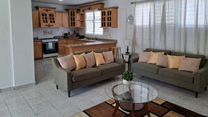 Una sala de estar moderna con dos sofás, una mesa de centro de vidrio y una cocina al fondo.