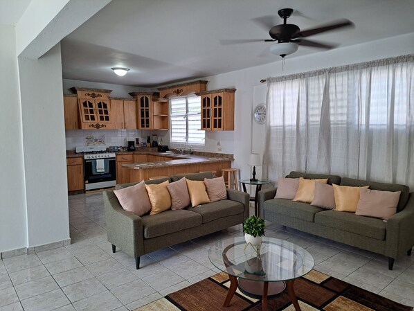 A modern living room with two sofas, a glass coffee table, and a kitchen in the background.