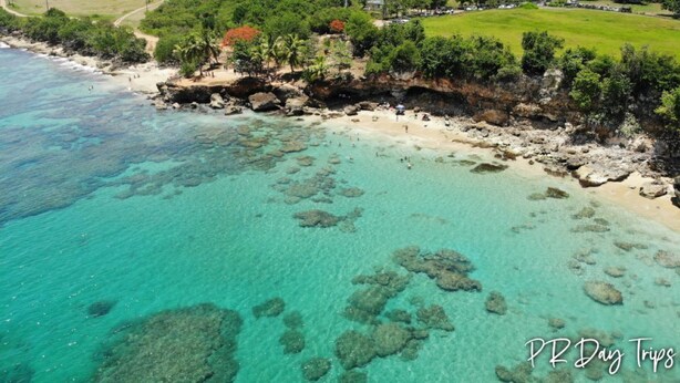 Vista aérea de una playa con agua turquesa cristalina, formaciones rocosas y una zona cubierta de césped y árboles.