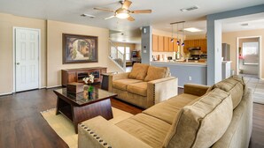 A spacious living room with a beige sofa, a glass coffee table, and a ceiling fan. A kitchen is visible in the background with wooden cabinets and a stainless steel refrigerator. - San Antonio Home w/ Private Hot Tub + Pool! (San Antonio)