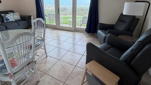 Una sala de estar con ventilador de techo, un sofá negro, una silla blanca, una mesita y vistas al exterior a través de puertas de cristal.