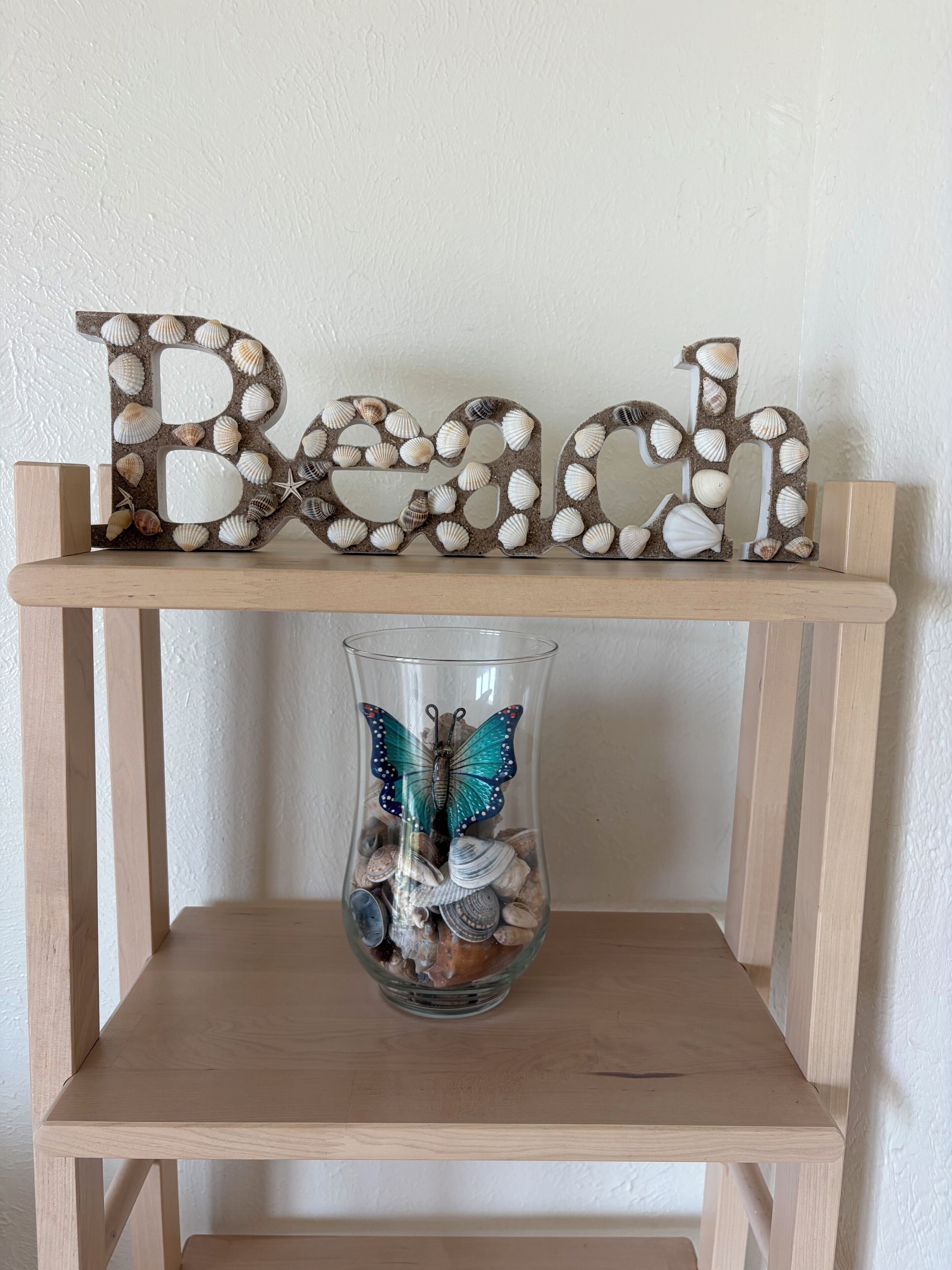 Een plank met een decoratief bordje met de tekst 'Beach' en een glazen vaas met schelpen en een vlinder.