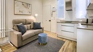 Un apartament modern amb un sofà beix, un otomà petit i rodó, una cuina amb armaris blancs i una finestra gran.