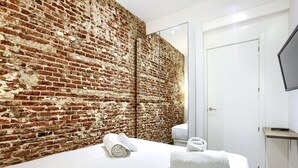 Una camera da letto moderna con parete in mattoni, un letto con lenzuola bianche, uno specchio e un televisore fissato alla parete.