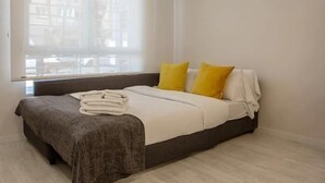 Ein ordentlich bezogenes Bett mit grauem Kopfteil, weißer Bettwäsche und zwei gelben Kissen.