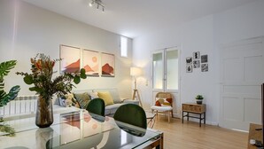 Ein modernes Wohnzimmer mit einem Glastisch, einer Couch und einem Sessel. An den Wänden befinden sich abstrakte Gemälde und auf dem Tisch steht eine Pflanze.