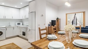 A modern kitchen with white cabinets, a microwave, and a washing machine. There is a dining area with a wooden table set for two, a TV, and a large mirror reflecting a window with curtains. - Plaza Mayor II - Two-bedroom apartment (Madrid)