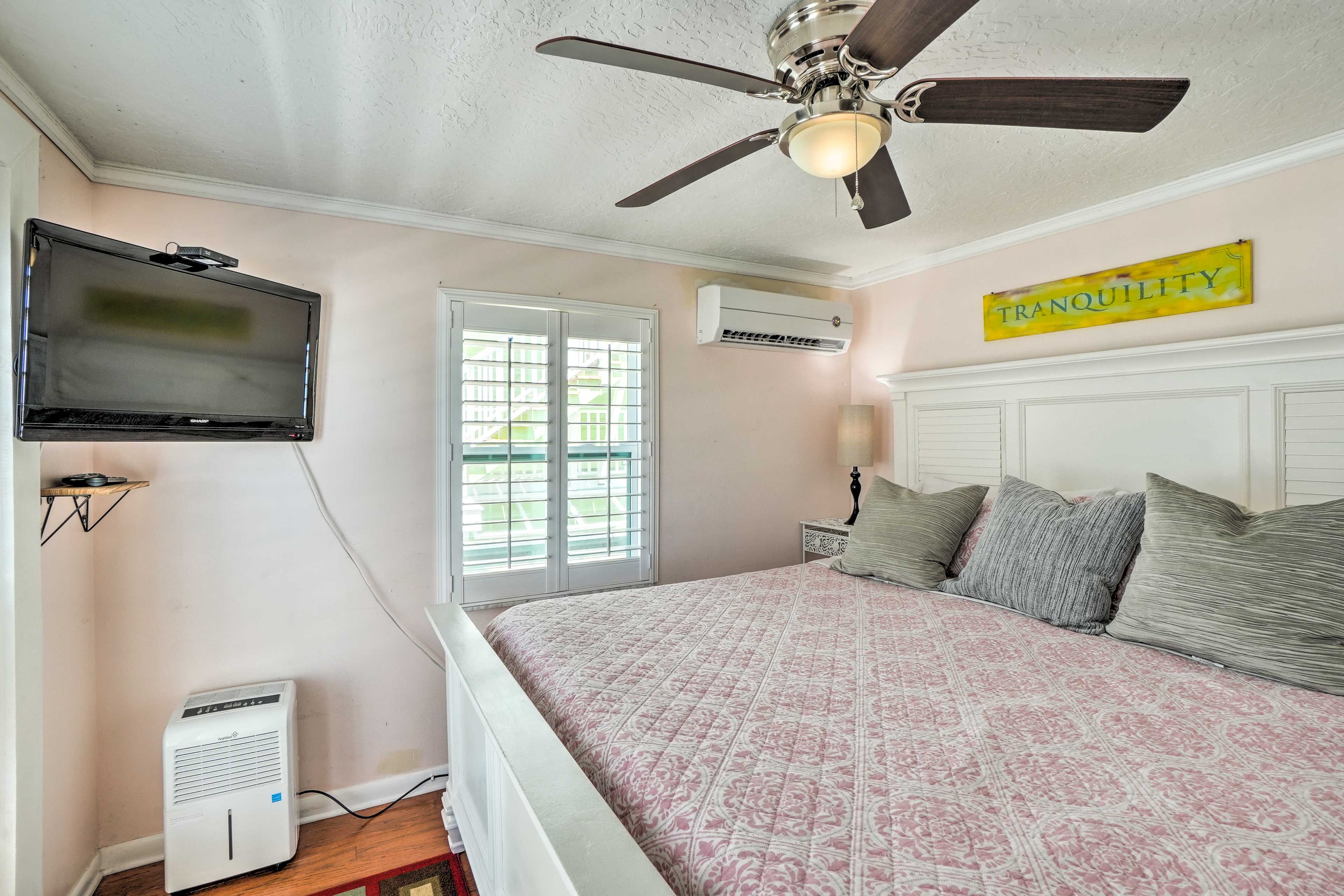 Un dormitorio con cama, ventilador de techo, televisión, aire acondicionado y ventana con persianas.
