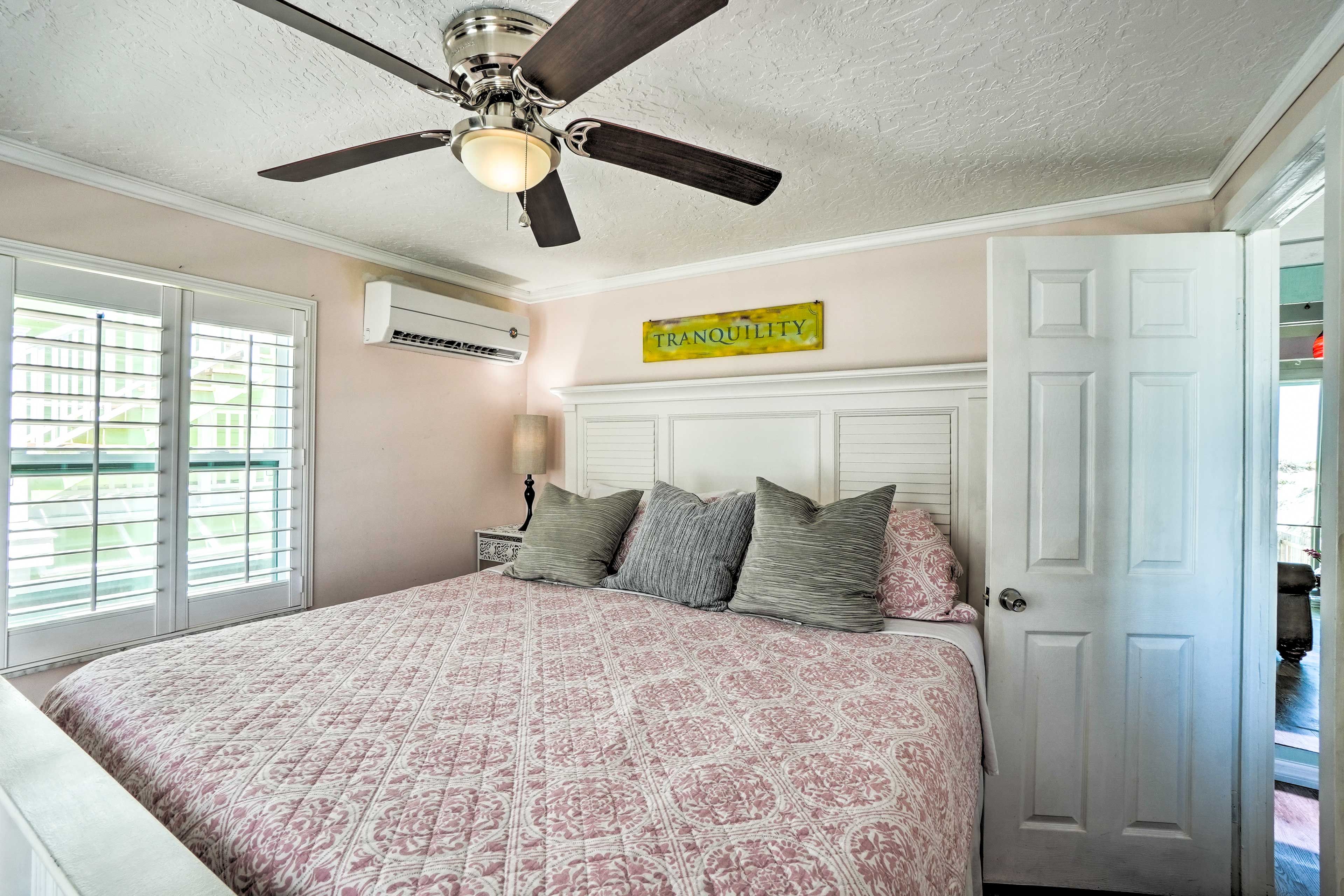 Un dormitorio con cama, ventilador de techo, ventana con persianas, unidad de aire acondicionado y una puerta.