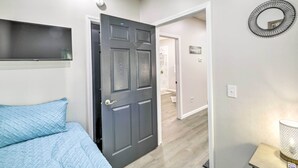 A bedroom with a bed, a dark door, a wall-mounted shelf, a round mirror, and a bedside table with a lamp.