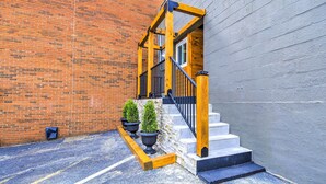 A brick building with a wooden staircase leading to a door, potted plants, and a parking area.