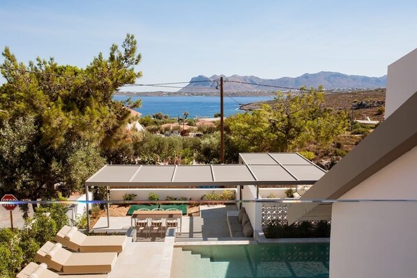 Ein Poolbereich mit Liegestühlen, einer überdachten Lounge und Blick auf das Meer und die Berge.