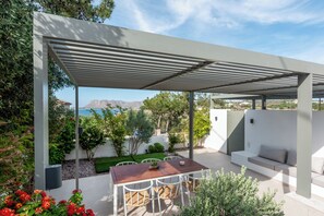 Eine überdachte Terrasse im Freien mit Tisch und Stühlen, einer Couch und Blick auf das Meer und die Berge.