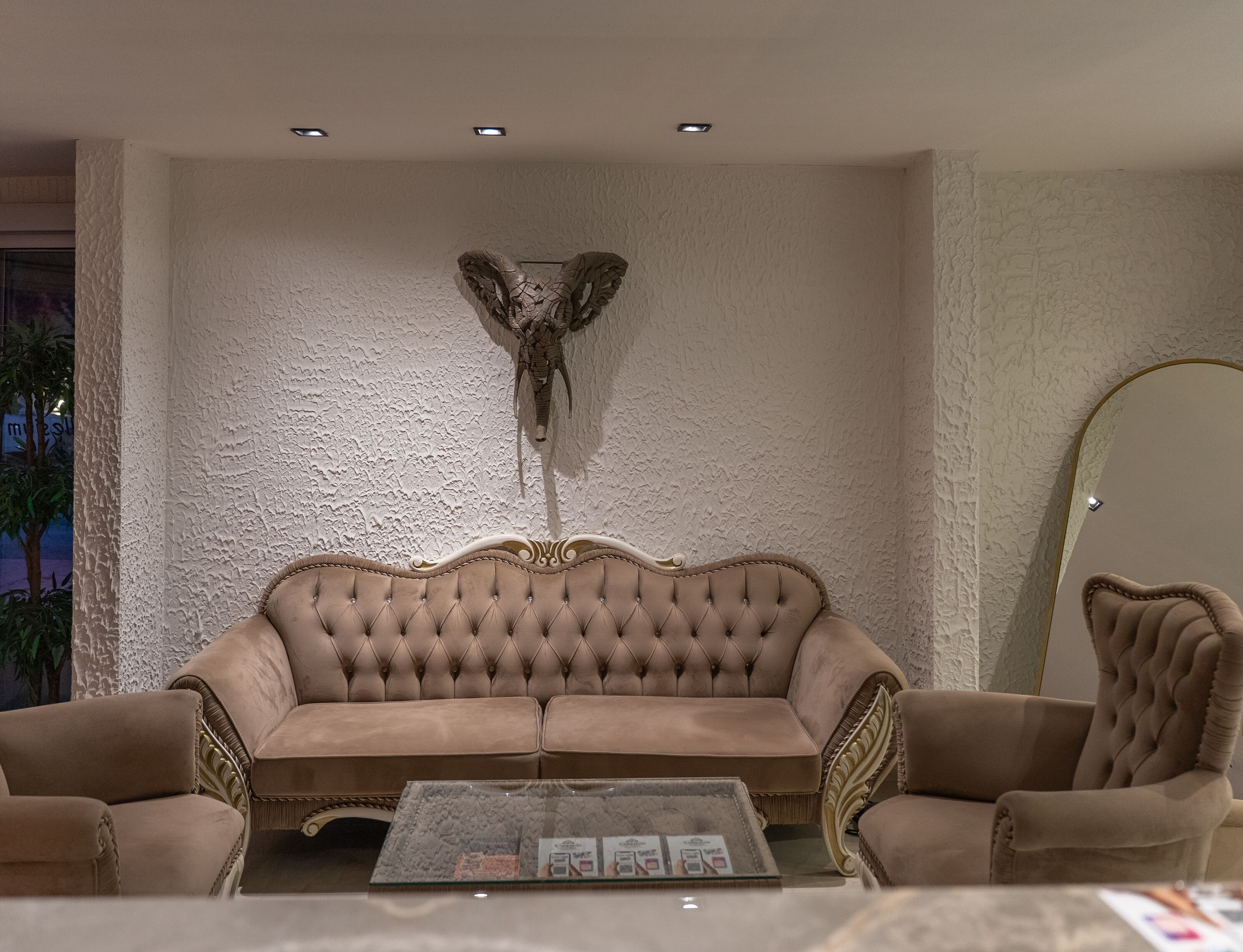 A living room with a tufted sofa, two armchairs, a glass coffee table, a decorative wall piece, and a round mirror.