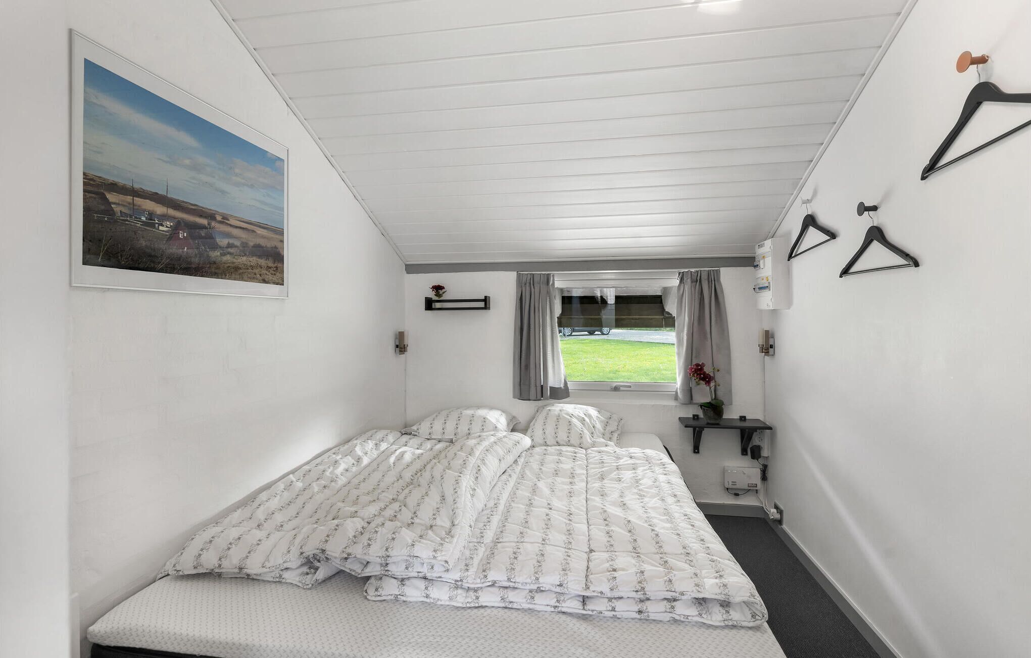 Un dormitorio con cama, ventana con cortinas, una mesita con un florero y un cuadro en la pared.