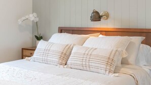 Un letto rifatto con biancheria bianca e cuscini decorati, una pianta sul comodino e un punto luce a parete.